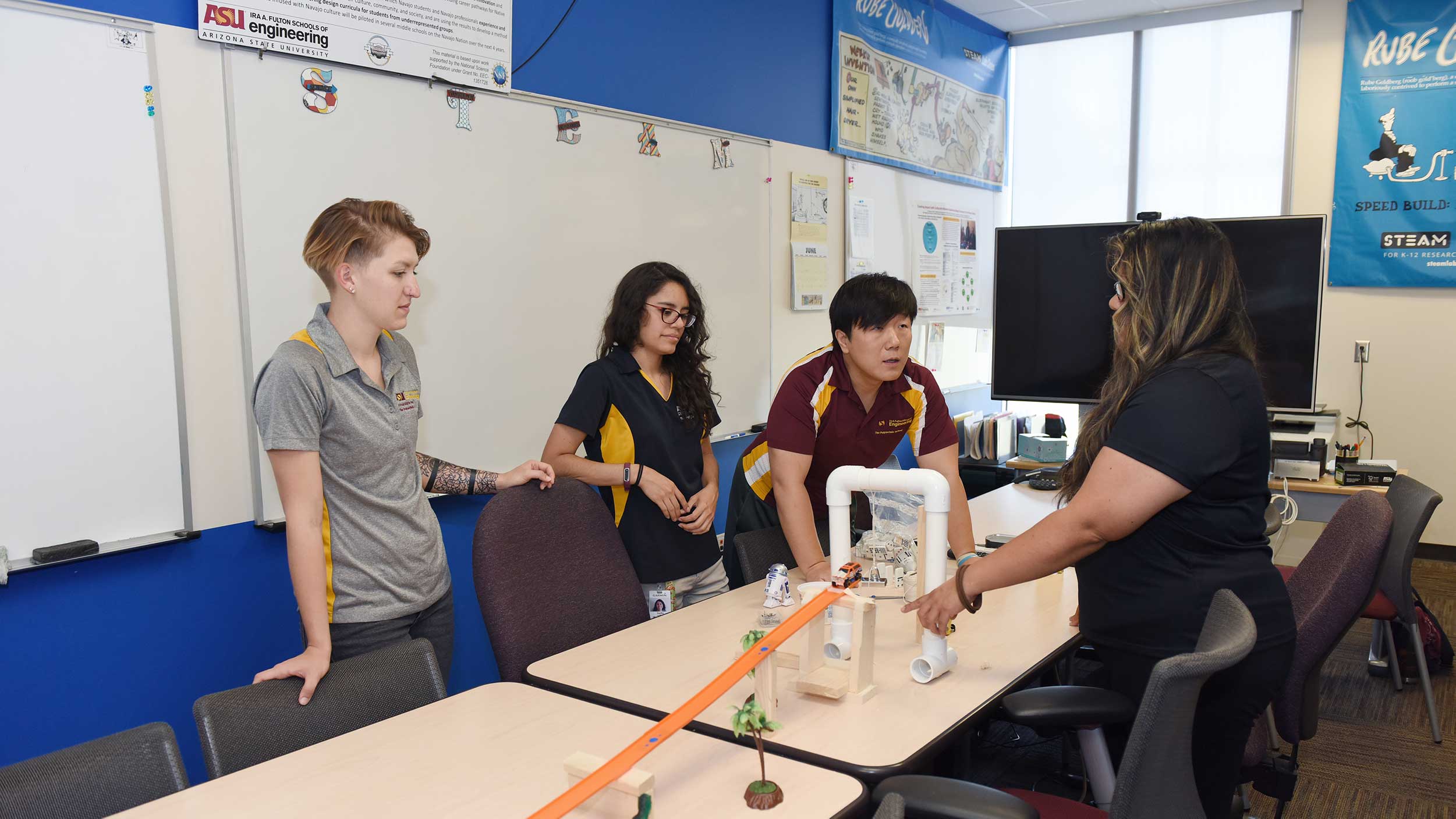 Four people stand around a table building a model of a structure as practice for how to teach engineering concepts.